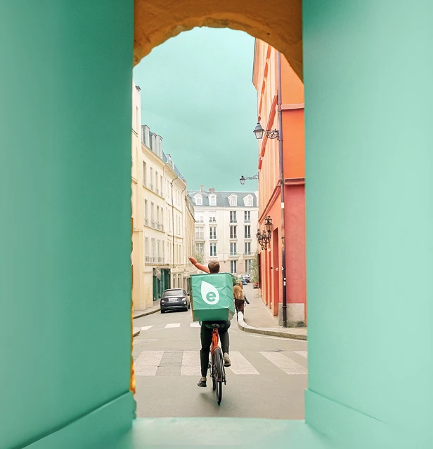 Vu à travers une arche turquoise et jaune, un coursier à vélo avec un grand sac à dos de livraison turquoise portant l'icône d'Ecolotrans, s'éloigne sur une rue de ville européenne.