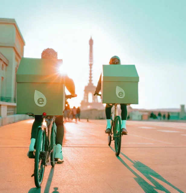 Deux coursiers à vélo avec des boîtes de livraison portant l'icône d'Ecolotrans, s'éloignent du spectateur en direction de la Tour Eiffel à Paris au coucher du soleil.